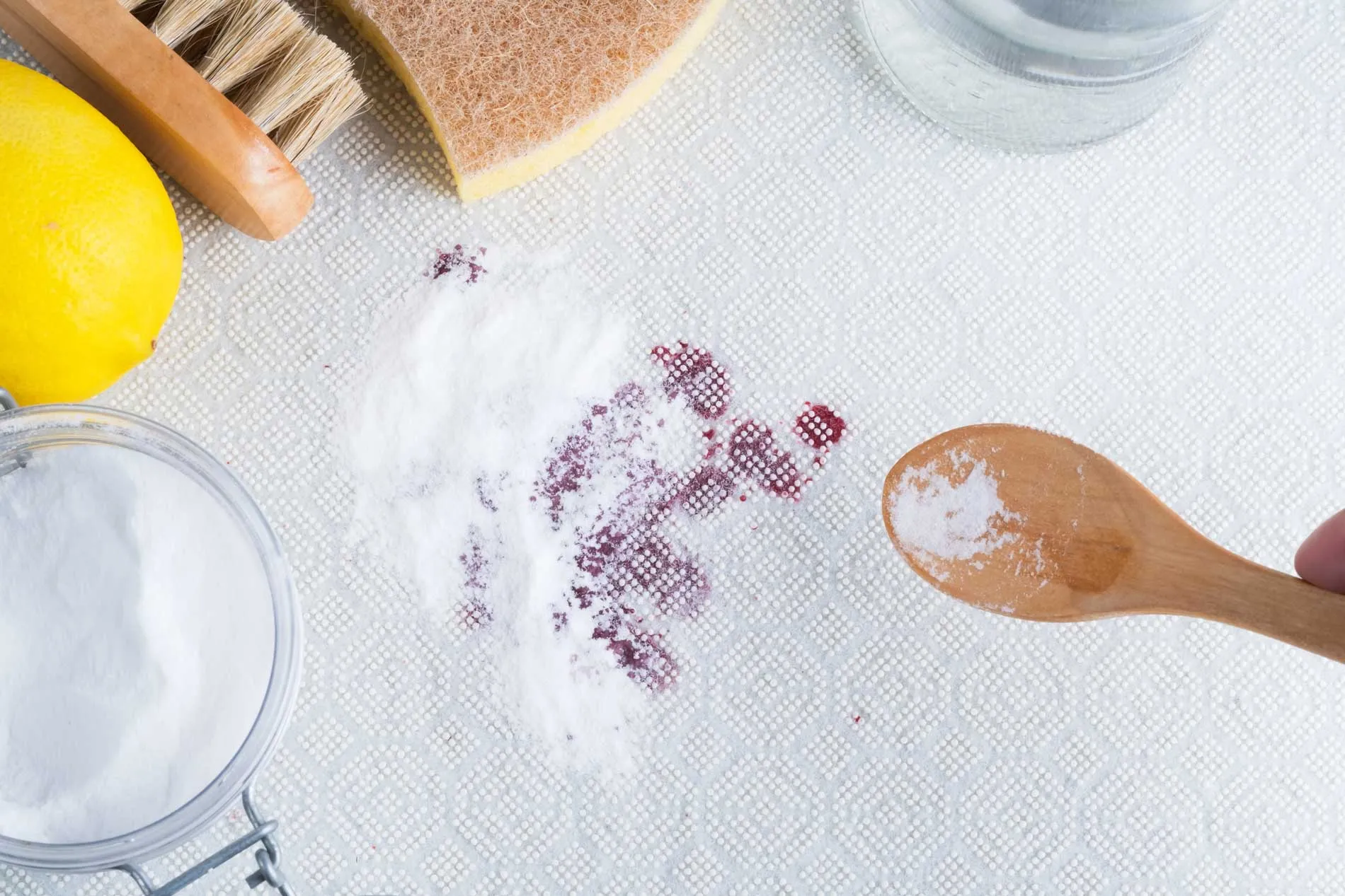 Baking soda spread over a red wine stain on a white rug, with a lemon and scrub brush nearby